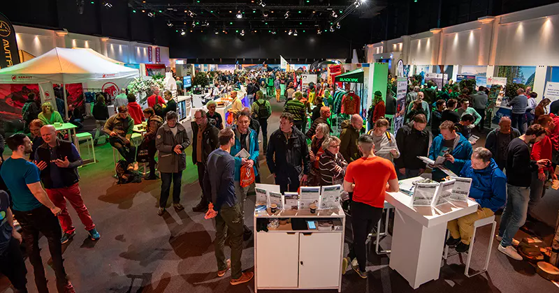 Een grote beurs in een overdekte ruimte waar veel mensen de stands bezoeken, met elkaar praten en producten bekijken onder felle verlichting, terwijl banners, displays en tenten zorgen voor een drukke, levendige sfeer.